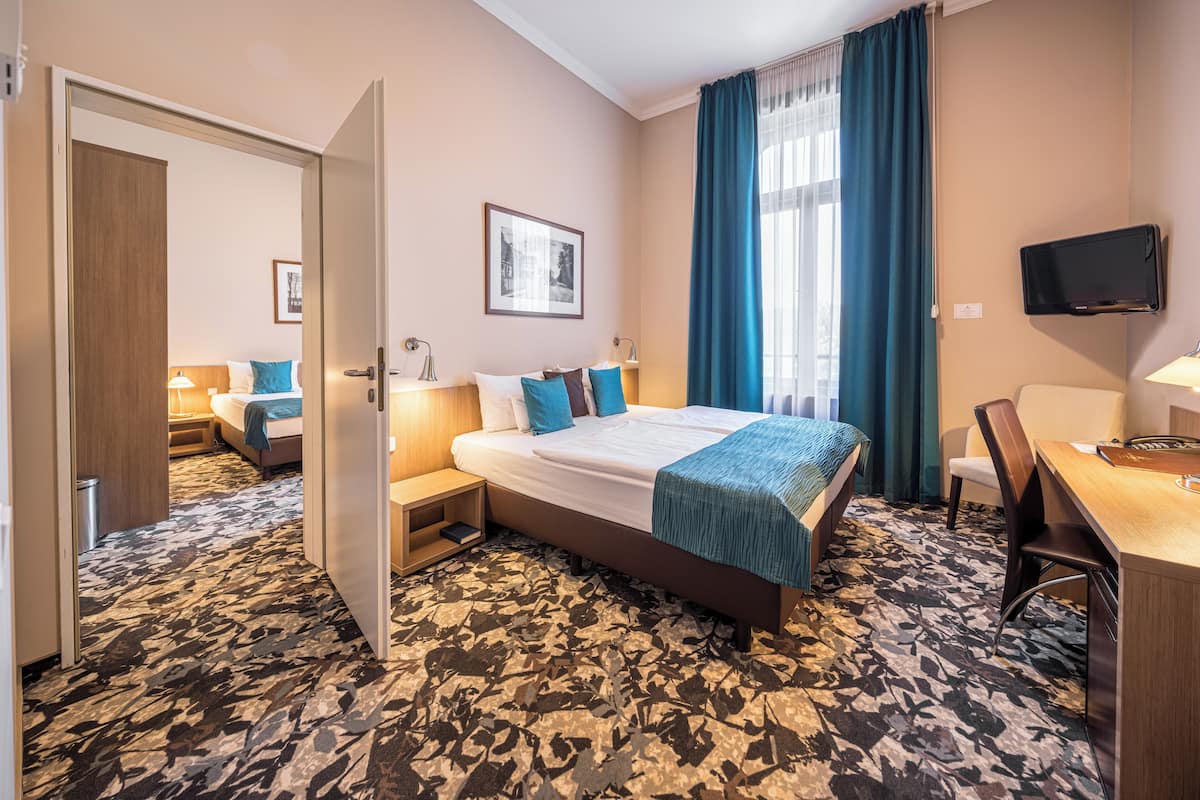 Habitación de hotel con una cama grande, un escritorio con silla, televisor montado en la pared y una ventana con cortinas.