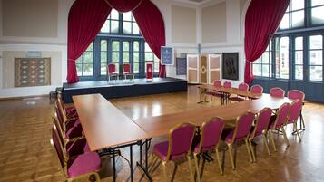 Un salón de conferencias con una larga mesa y sillas, ventanas amplias con cortinas rojas y un escenario.