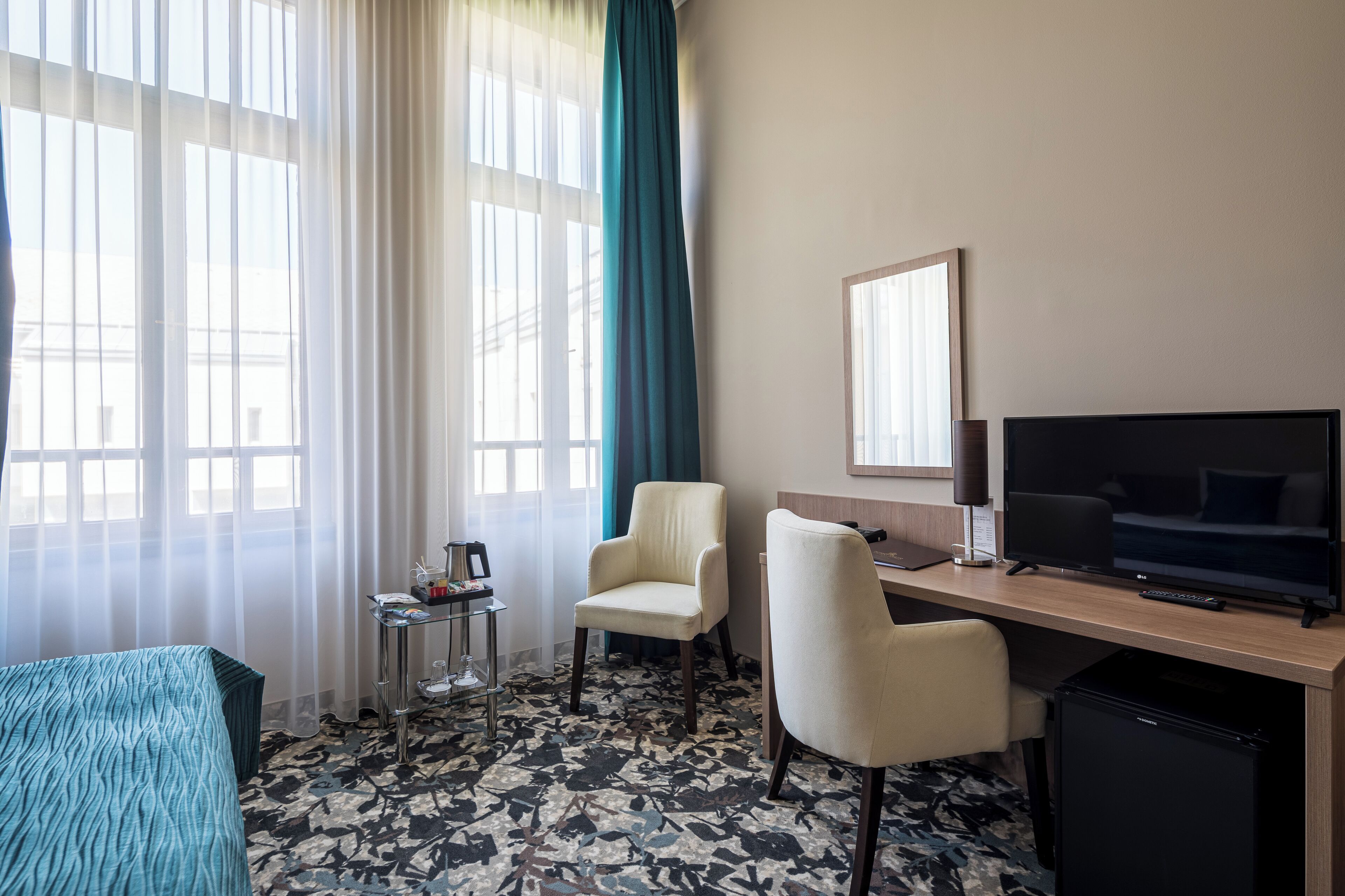 Een hotelkamer met een bed, een bureau met televisie, een stoel, een klein tafeltje met een lamp en een minibar.