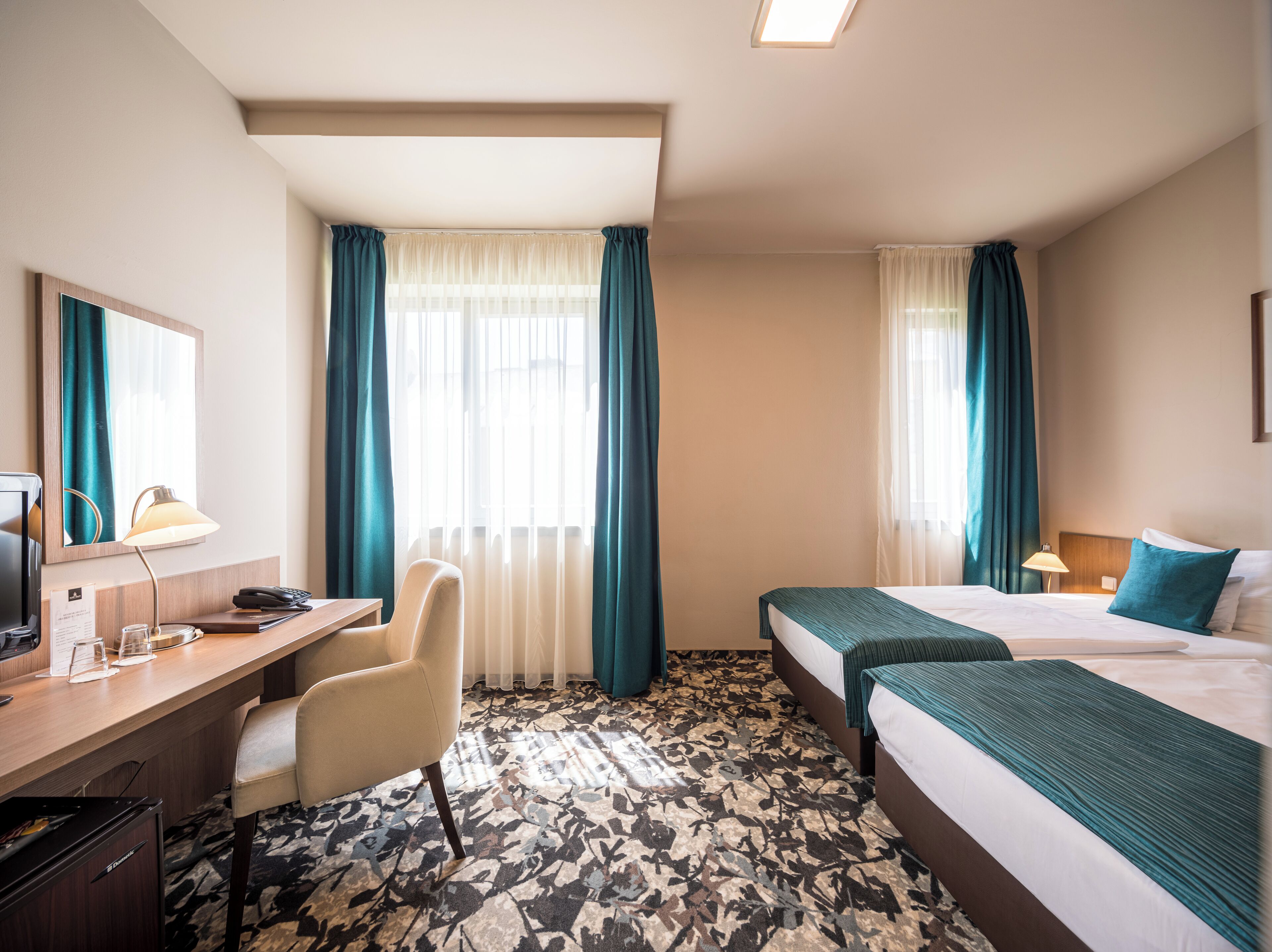 A hotel room with a desk, chair, two beds, and a large window with curtains.