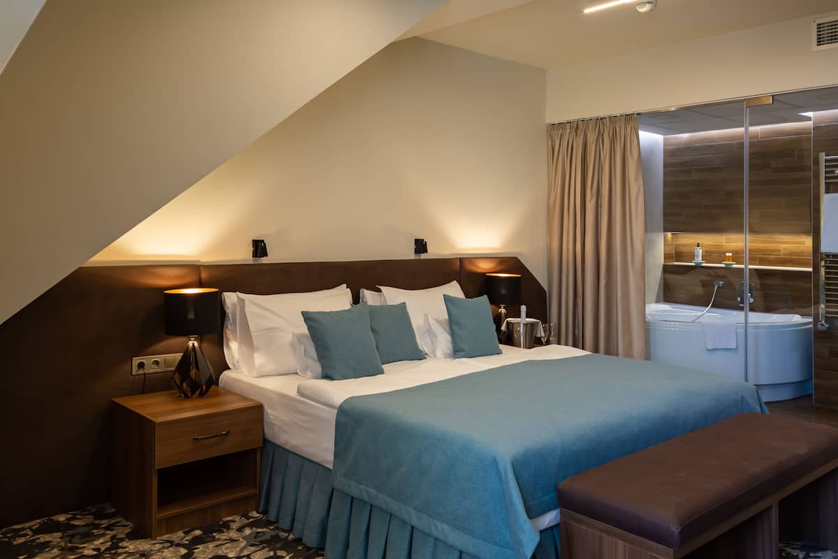 Una habitación de hotel moderna con una cama grande, lámparas de noche, una mesita de noche, un banco y un baño separado con bañera y ducha.