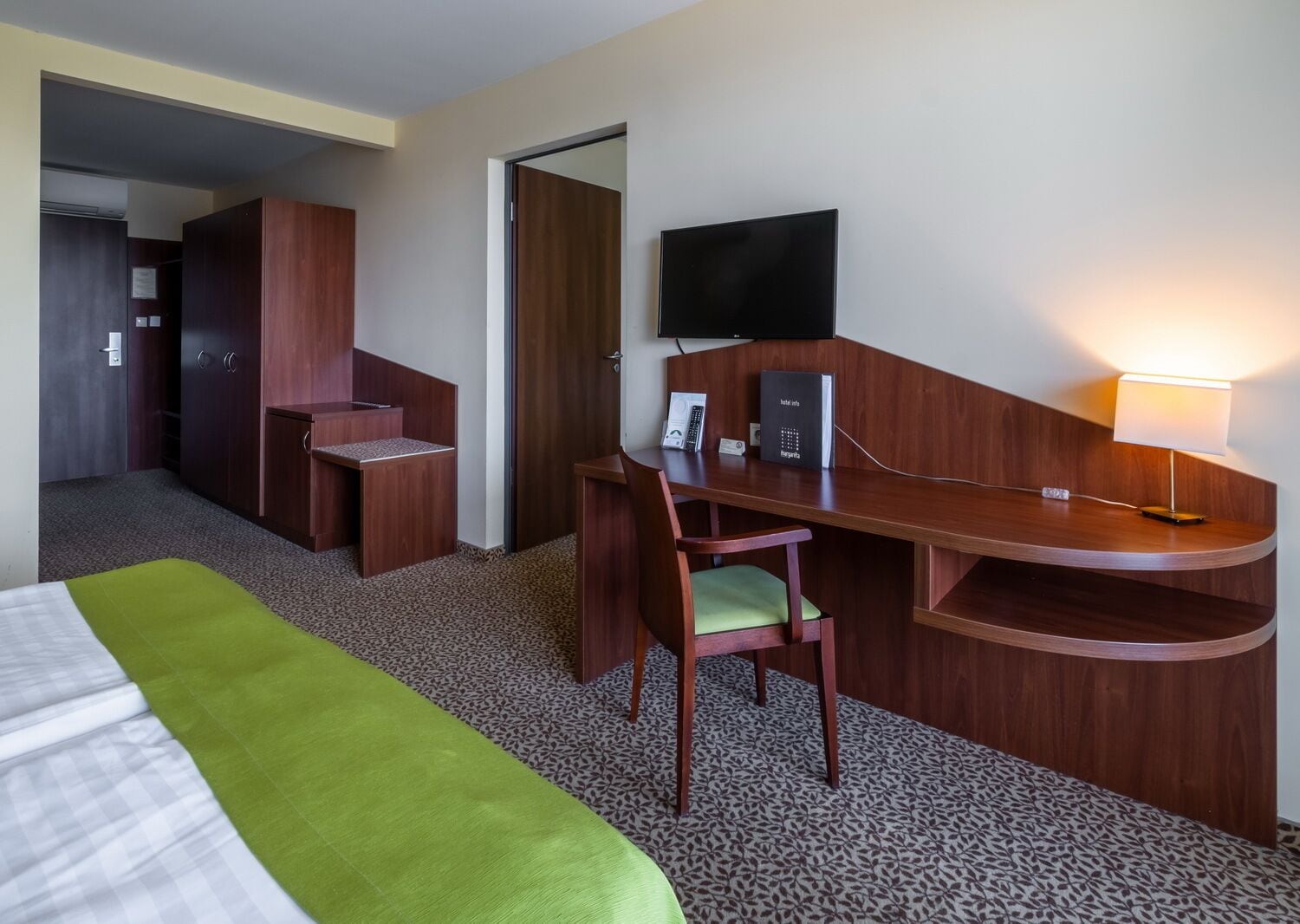 A hotel room with a bed, a desk with a chair, a flat-screen TV, a telephone, and a lamp.