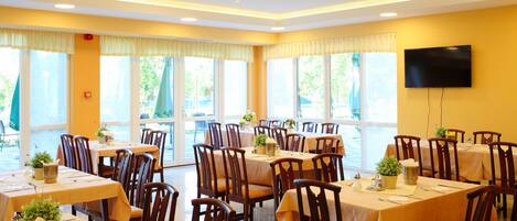 A dining area with multiple tables and chairs, large windows, and a chandelier.