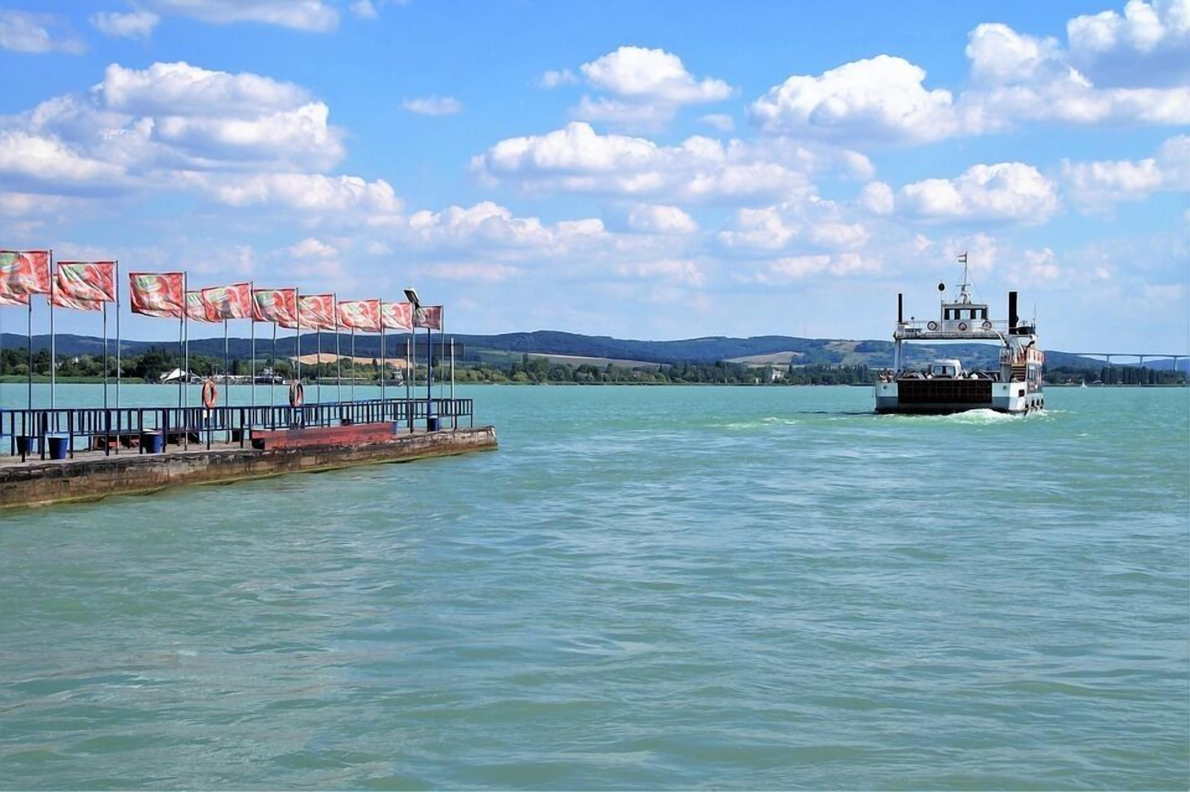 Un traversier traverse un lac paisible, avec un quai sur la gauche et un pont en arrière-plan.