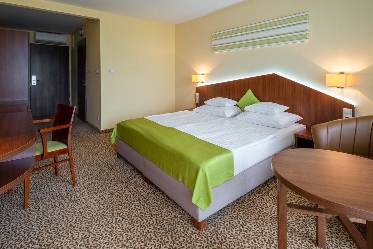 Une chambre d’hôtel avec un lit, deux lampes de chevet, une tête de lit en bois, un plaid vert, une moquette à motifs et un bureau avec une chaise.