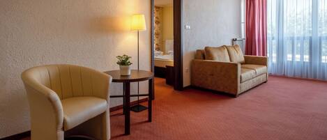 A hotel room with a beige armchair, a sofa, a small table with a plant, and a lamp.
