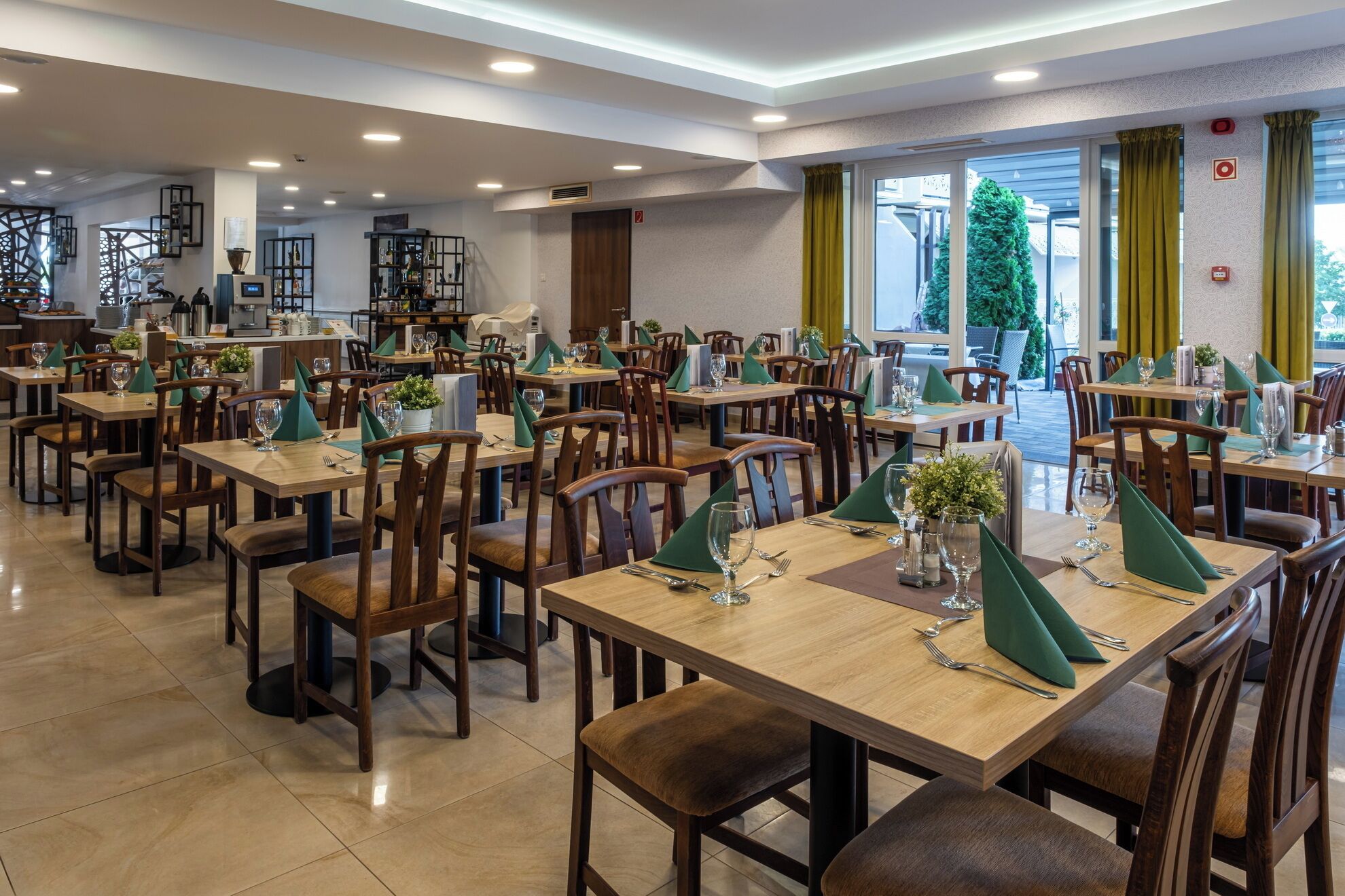Un comedor amplio con varias mesas y sillas, una cocina al fondo y ventanas grandes con cortinas.