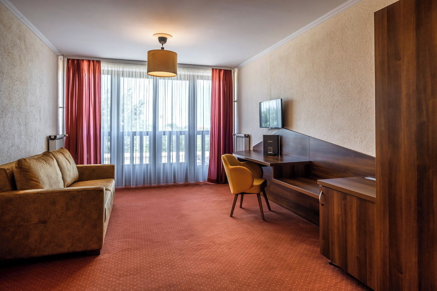 A hotel room with a sofa, a desk, a chair, a television, and a large window with curtains.