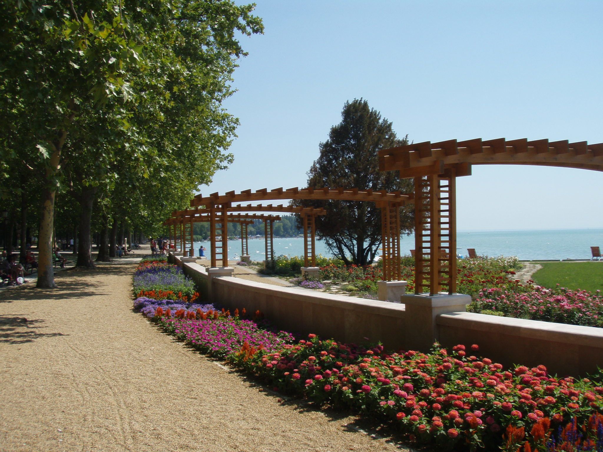 A pathway lined with flower beds and wooden pergolas overlooking a body of water.