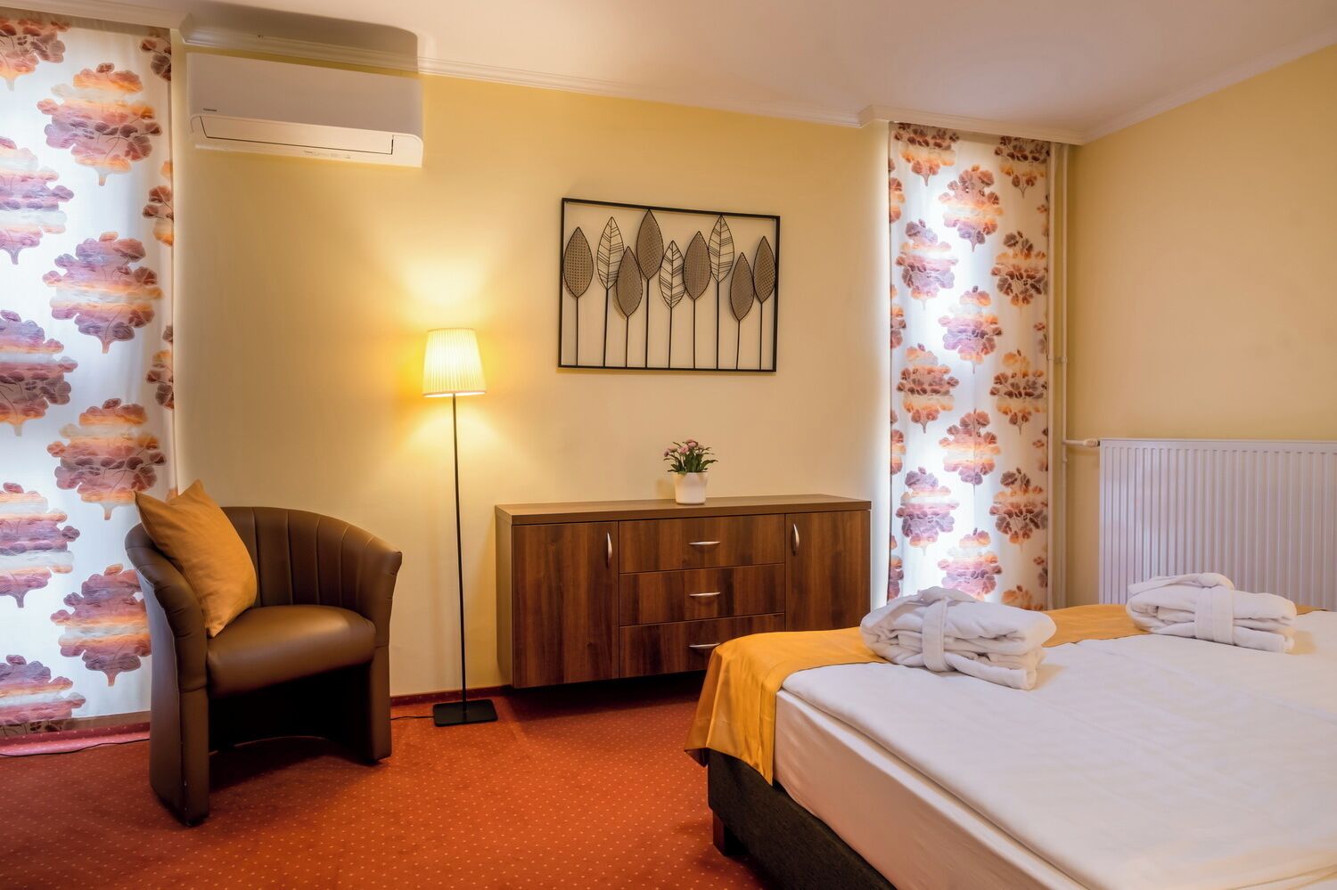 Ein Hotelzimmer mit Bett, Sessel, Schrank, Lampe und blumigem Wandpapier.