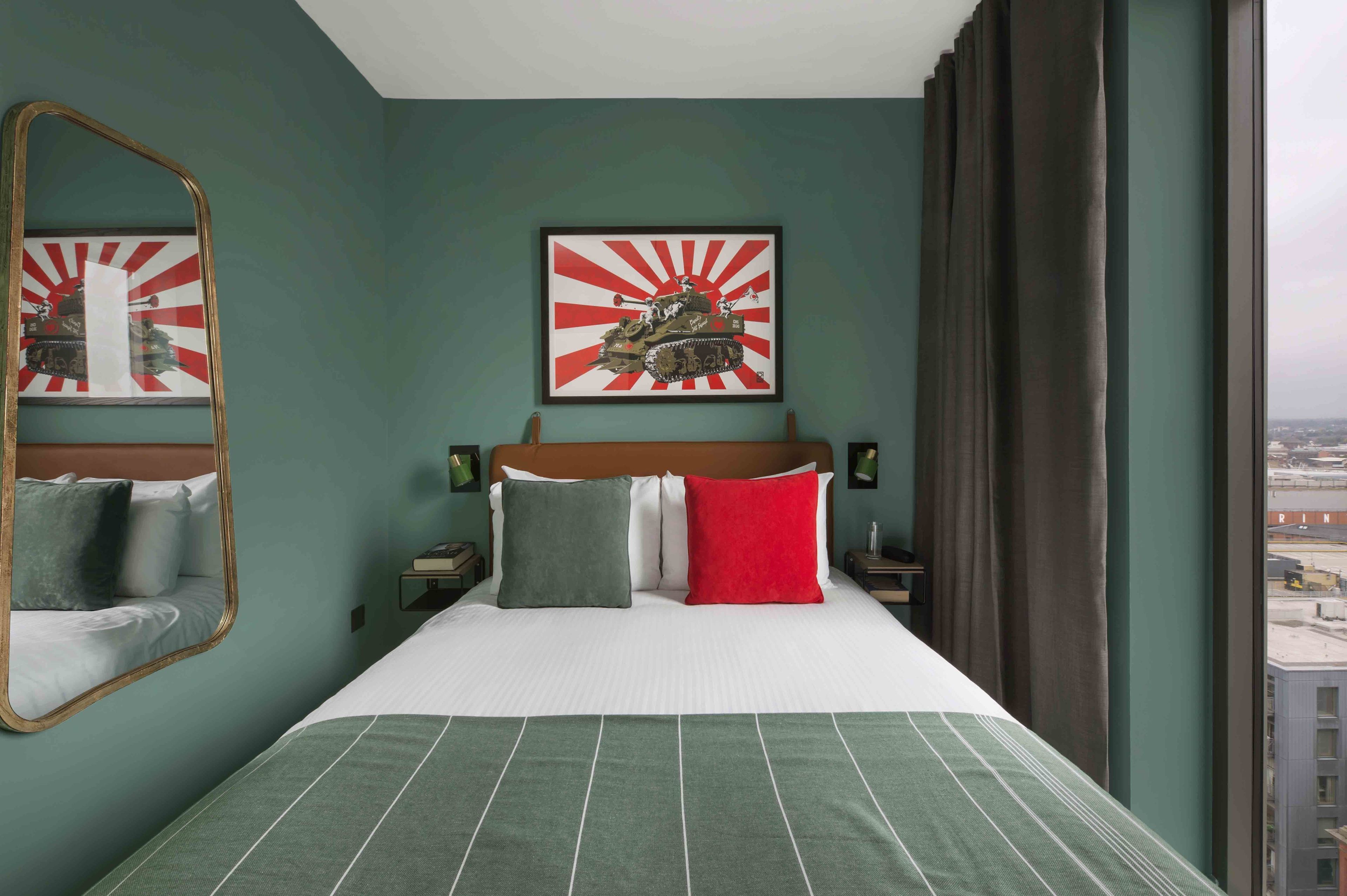 A hotel room with a bed, a large mirror, a framed picture of a tank, and a view of the city through the window.