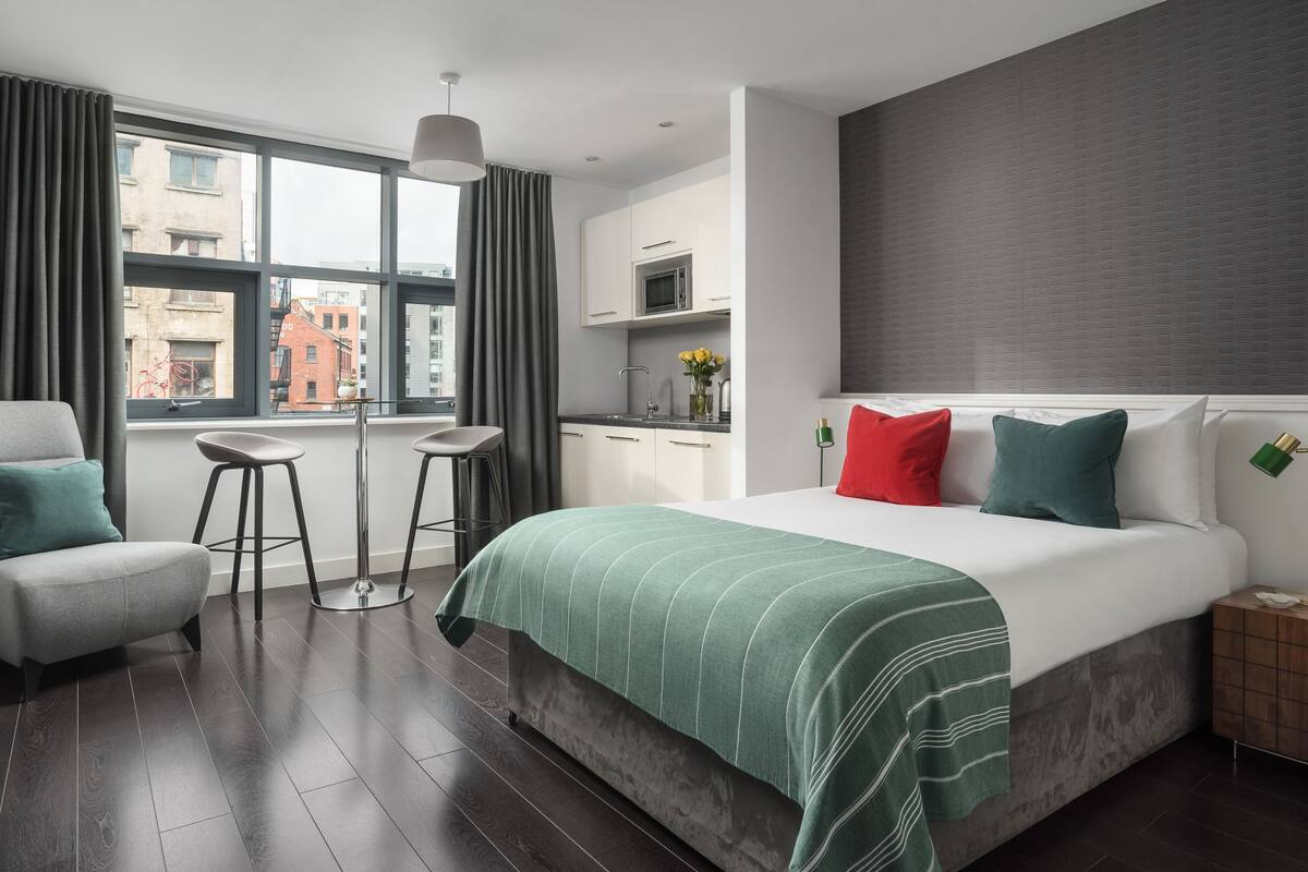 Habitación de hotel moderna con una cama grande, una pequeña cocina y vistas a la ciudad a través de amplios ventanales.