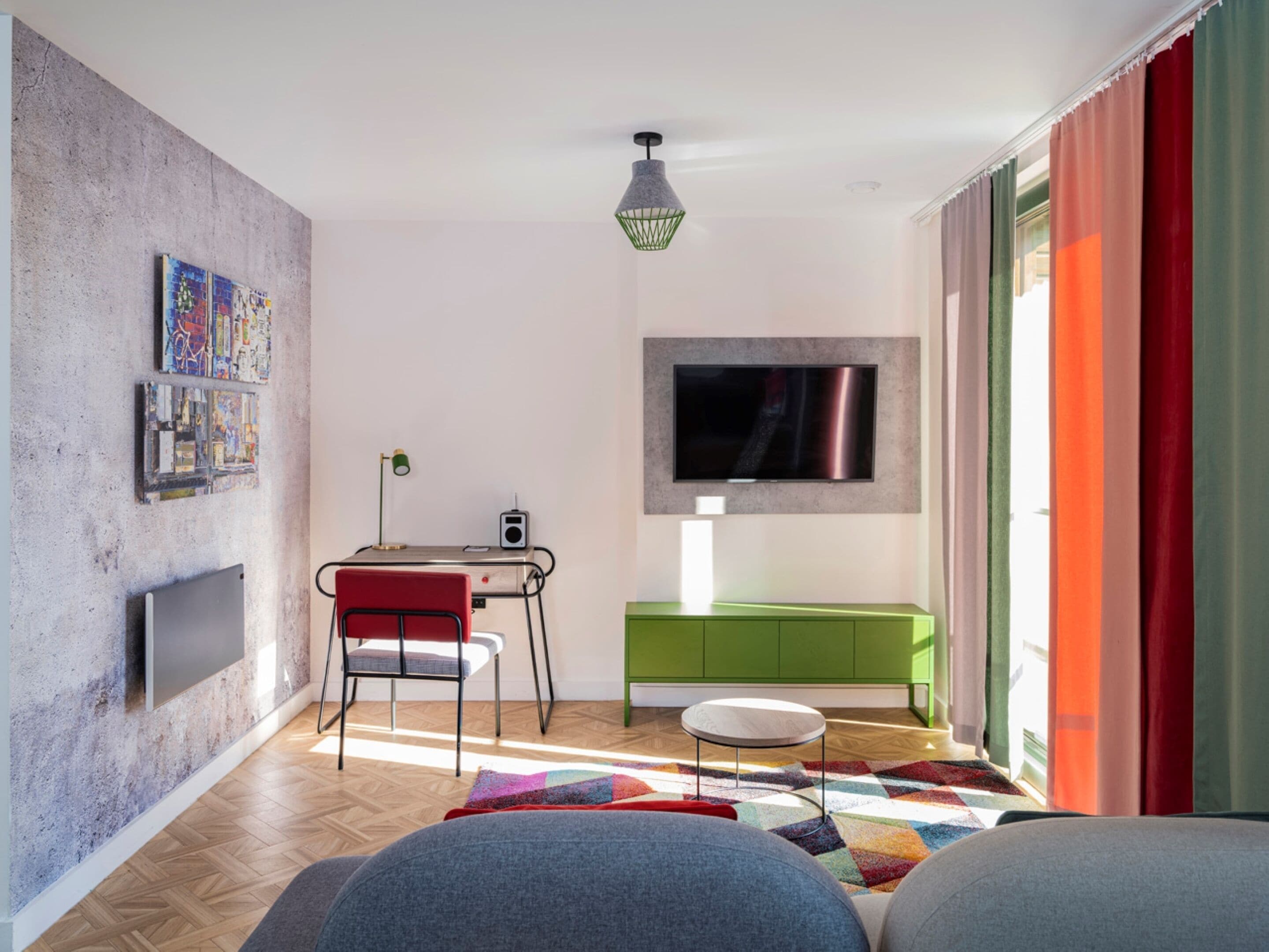 Un salón moderno con televisor de pantalla plana, un escritorio con silla, un armario verde y una alfombra colorida.