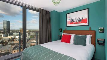 A hotel room with a large bed, teal walls, a city view, and a framed artwork above the bed.