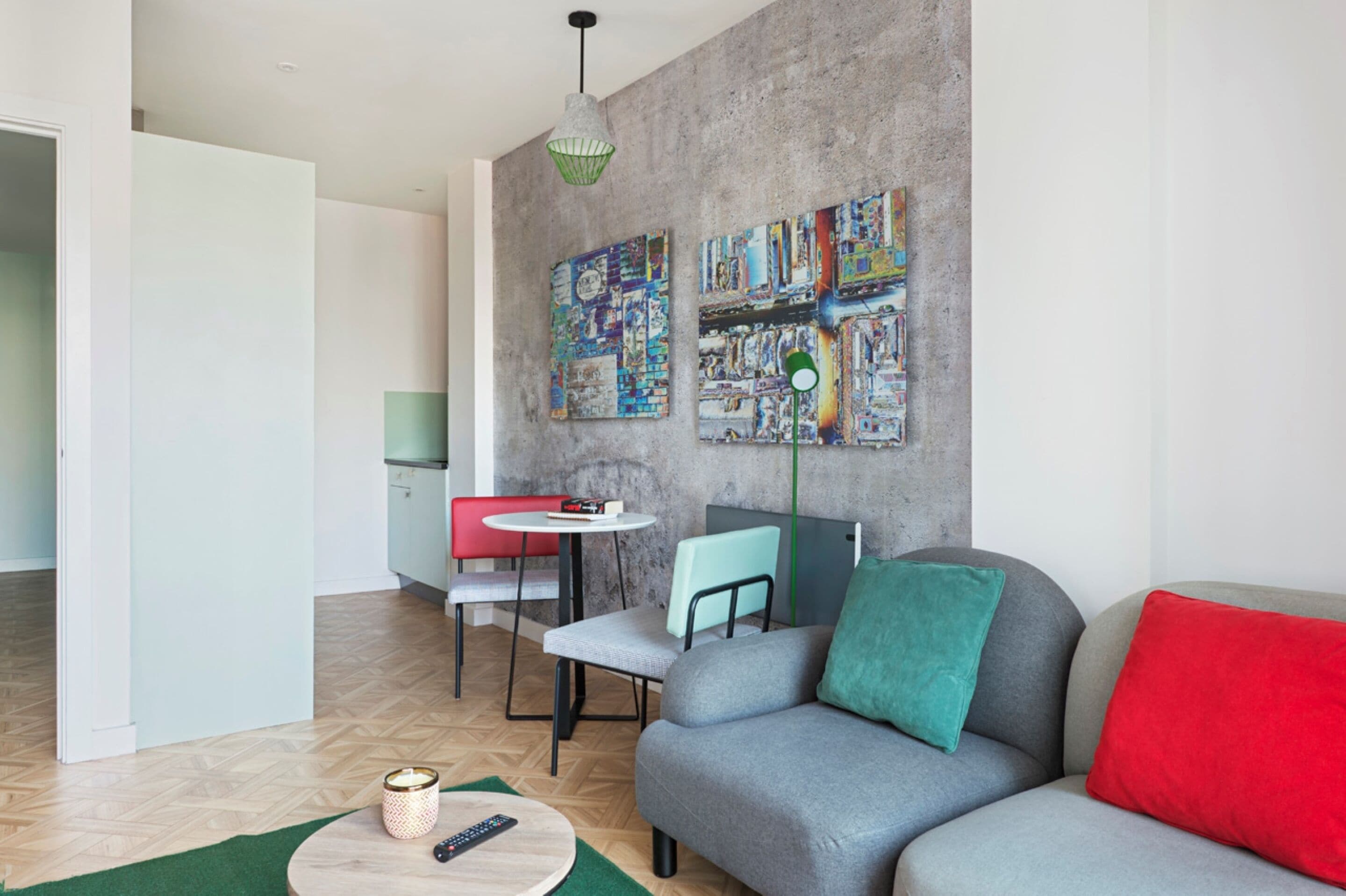 A modern living room with a grey sofa, red and teal cushions, a small round table, and a red chair. There are two colorful paintings on the wall and a green pendant light.