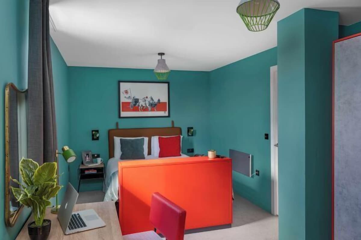 Un dormitorio con cama, escritorio, silla, planta y un cuadro en la pared.