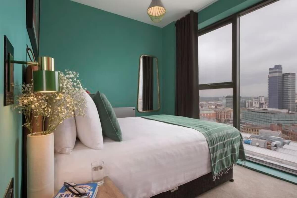 Un dormitorio con cama, ventana con vista a la ciudad, mesita de noche con lámpara y un jarrón con flores.