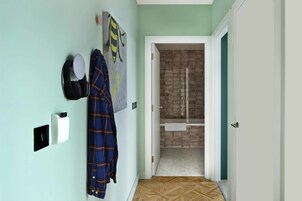 Un pasillo con pared verde claro, una chaqueta azul de cuadros colgada en la pared, un perchero blanco y un baño visible a través de una puerta abierta.