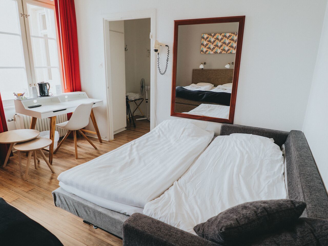 A room with a bed, a desk, and a chair. There is a mirror on the wall and a window with curtains.