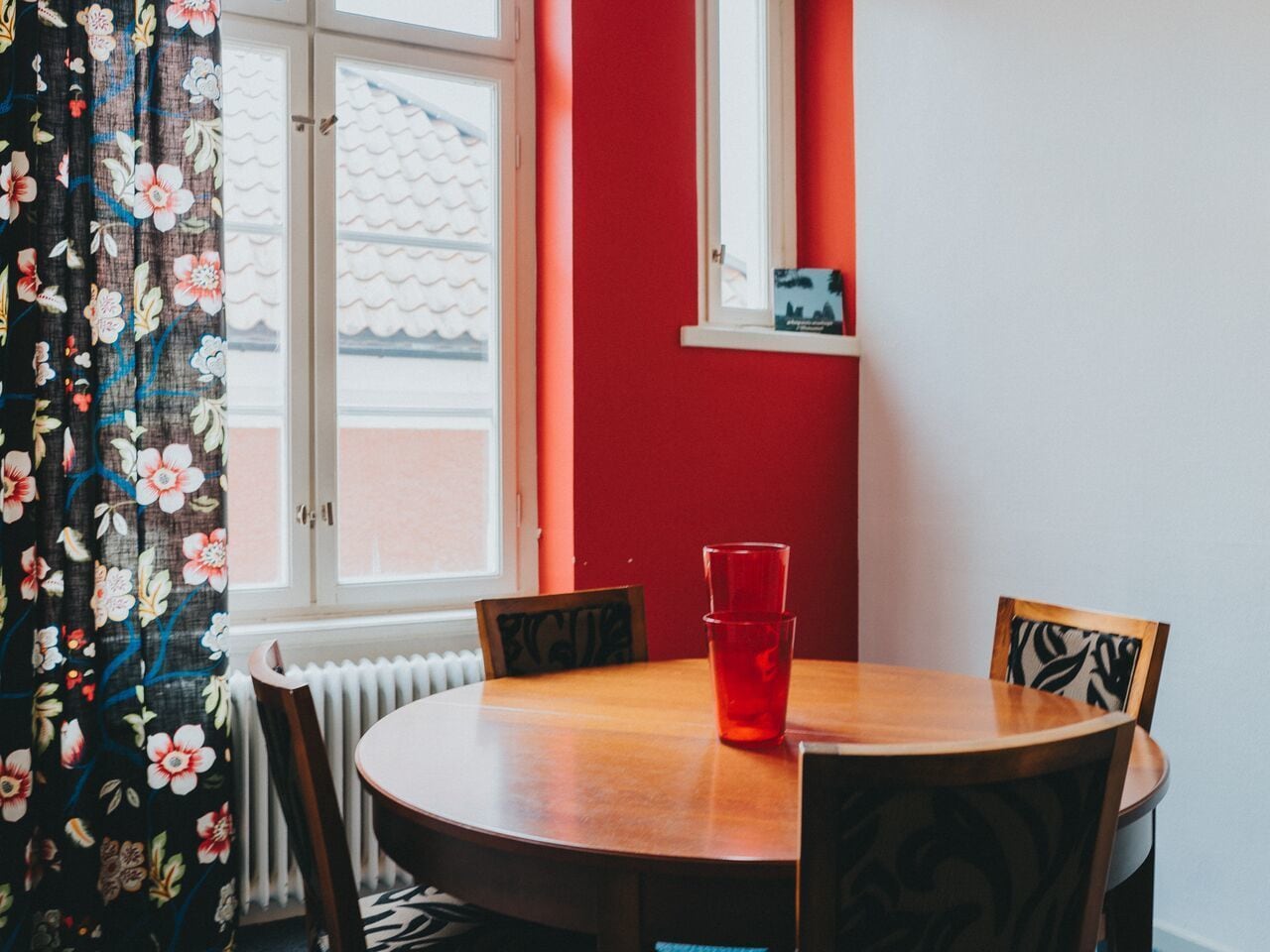A dining area with a round wooden table, two chairs, a red wall, a window with white shutters, and a red cup on the table.