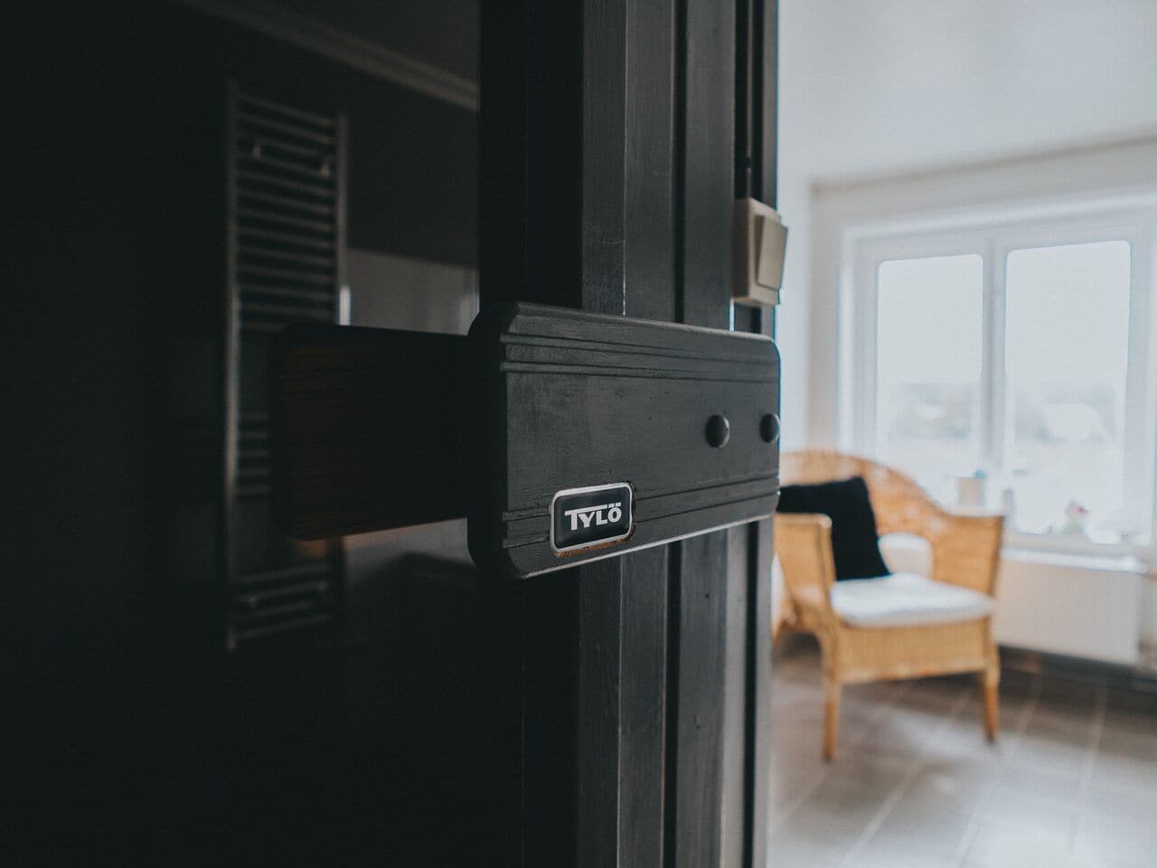A black metal door with a "TYLO" logo, a window with blinds, and a wicker chair with a cushion in a room with a white floor.