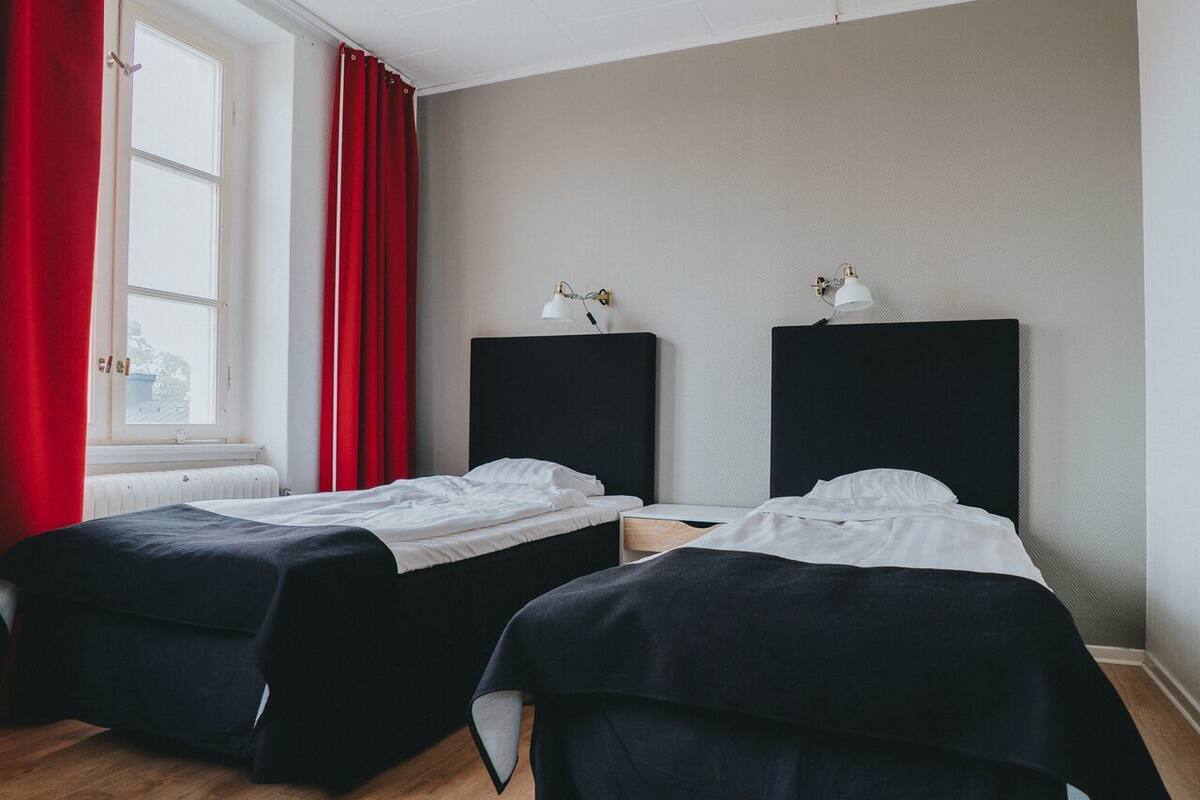A room with two beds, a window with red curtains, and two wall-mounted lamps.
