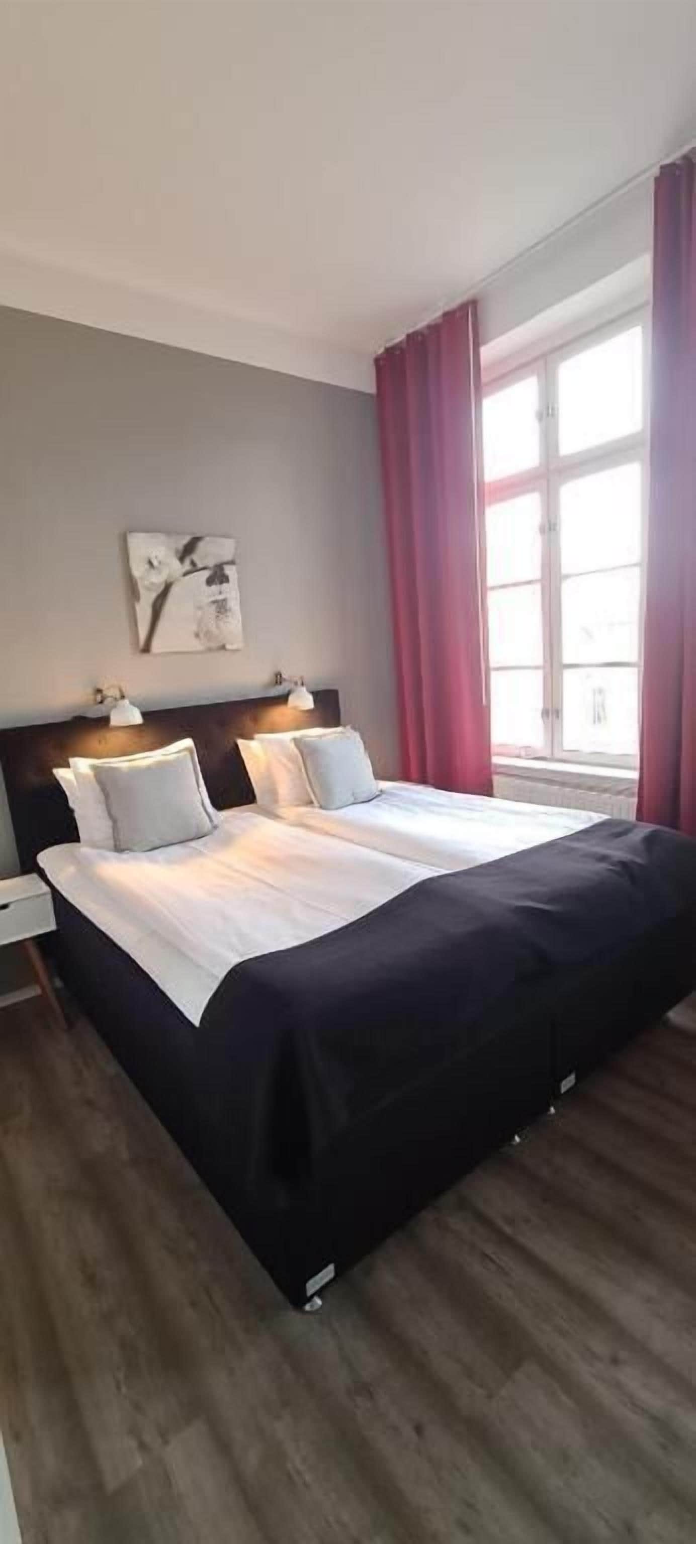 Ein Doppelbett mit dunklem Kopfteil, zwei Nachttische mit Lampen, ein großes Fenster mit roten Vorhängen und ein an der Wand befestigtes Kunstwerk.