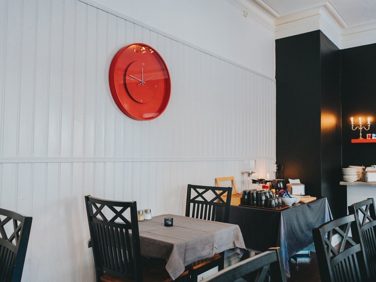 Uma área de refeições com um relógio vermelho redondo na parede, uma mesa com toalha de mesa cinza e cadeiras pretas com design em cruz.