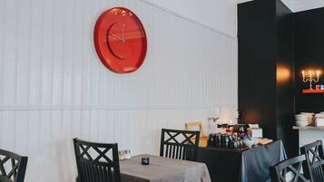 Uma área de refeições com um relógio vermelho redondo na parede, uma mesa com toalha de mesa cinza e cadeiras pretas com design em cruz.