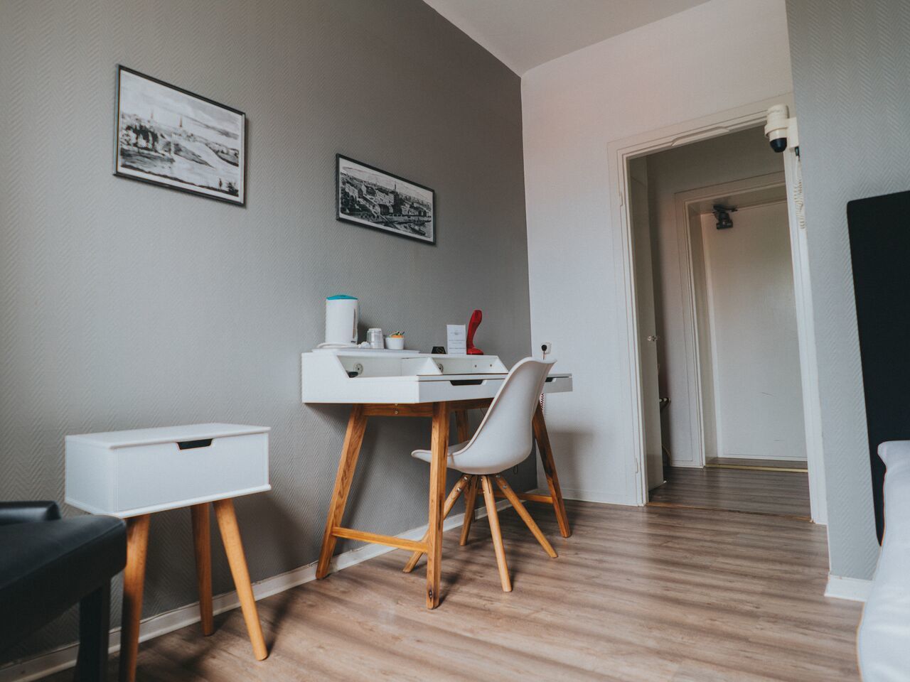 A modern room with a white desk, chair, and storage unit. There are two framed pictures on the wall and a door leading to another room.