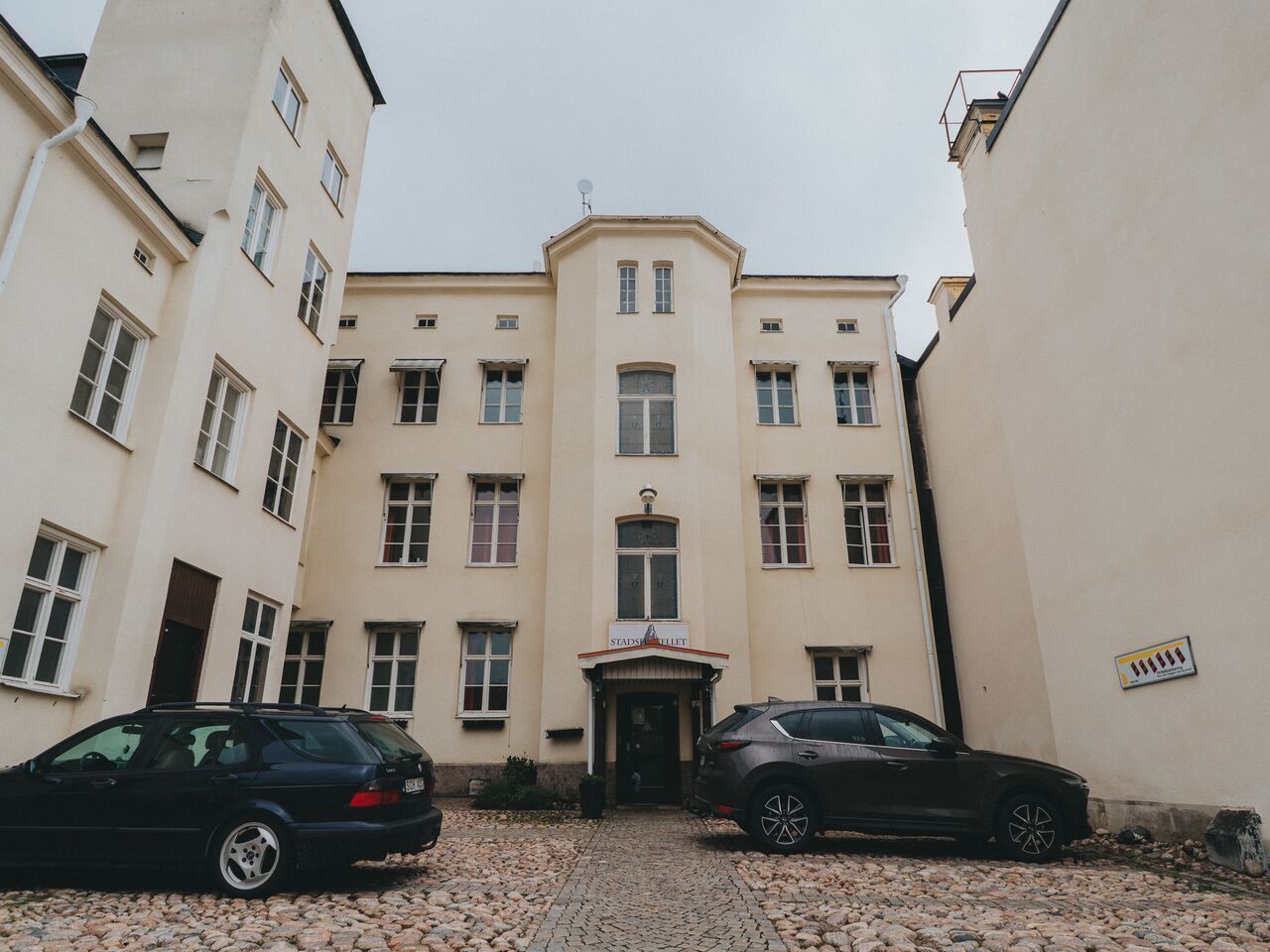 Un edificio con entrada central, dos coches aparcados y suelo adoquinado.