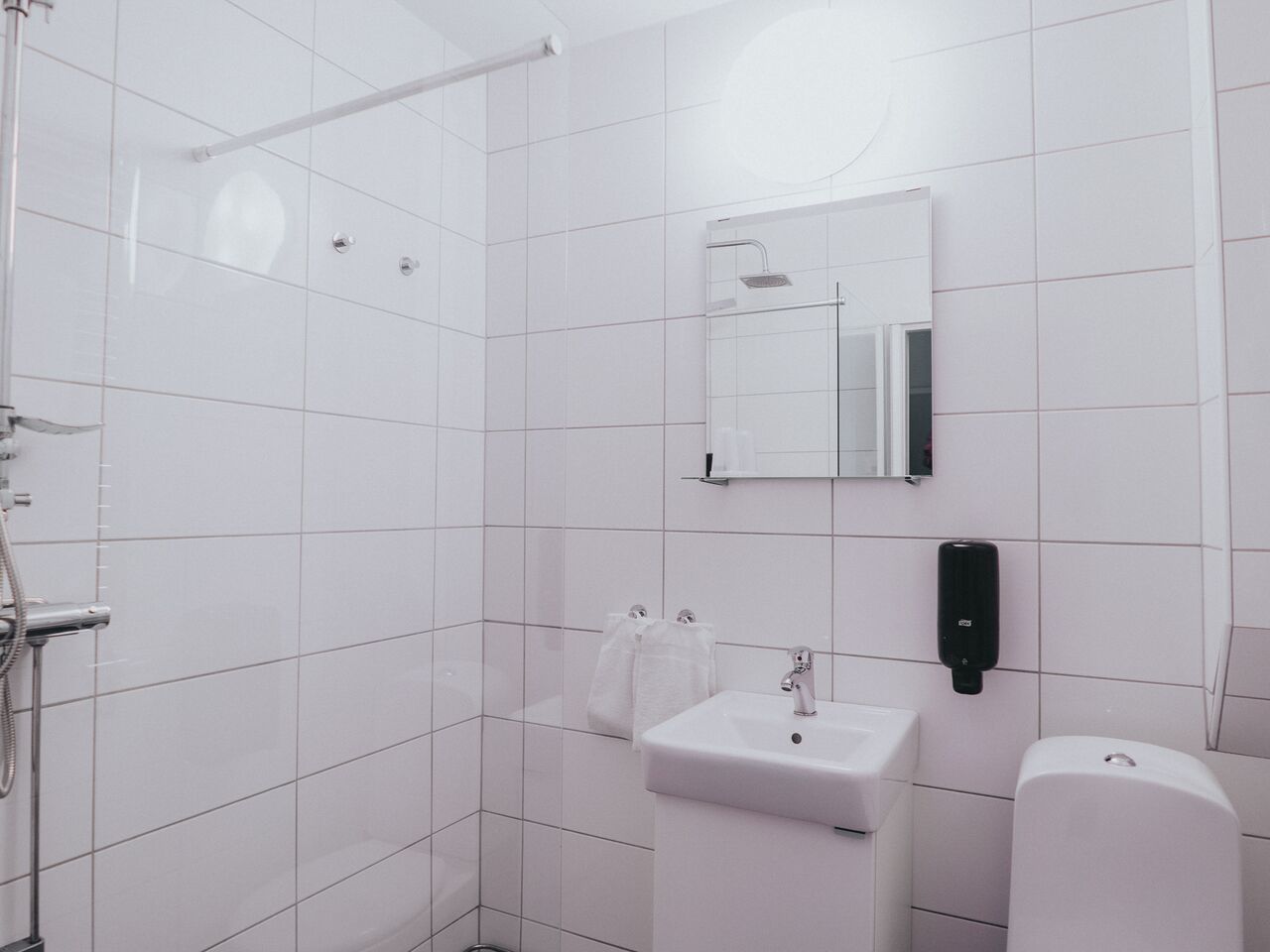 Un baño moderno con azulejos blancos, cabina de ducha con mampara de cristal, lavabo, espejo y dispensador de jabón.