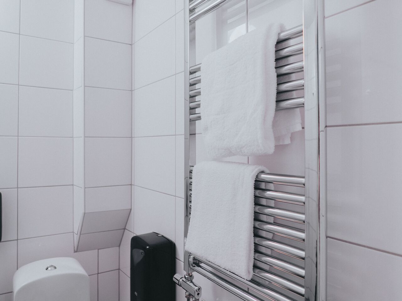 Een badkamer met witte tegels, een handdoekrek met handdoeken en een zwarte zeepdispenser.
