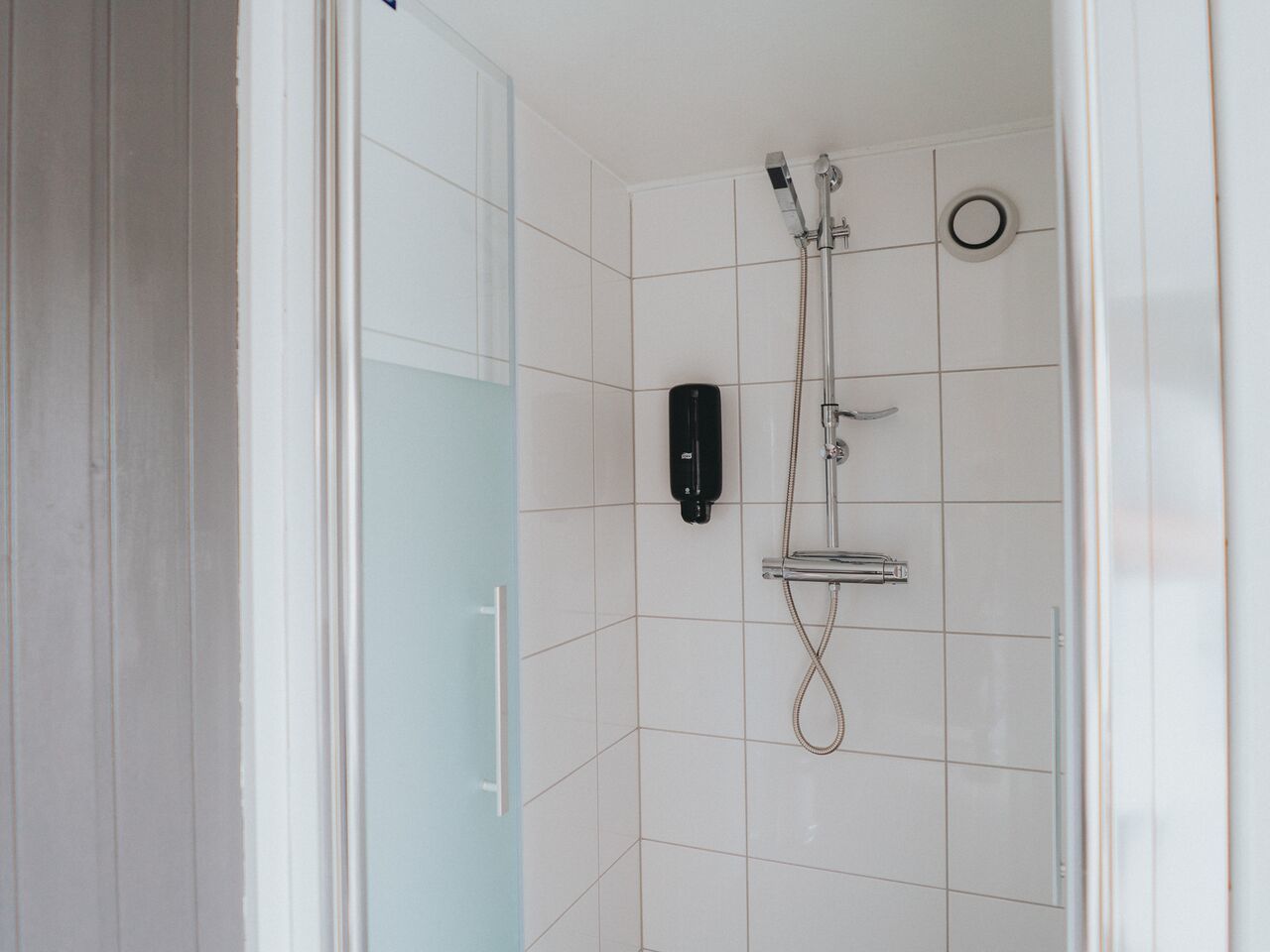 Un baño moderno con azulejos blancos, mampara de vidrio, cabezal de ducha de mano y grifo empotrado.