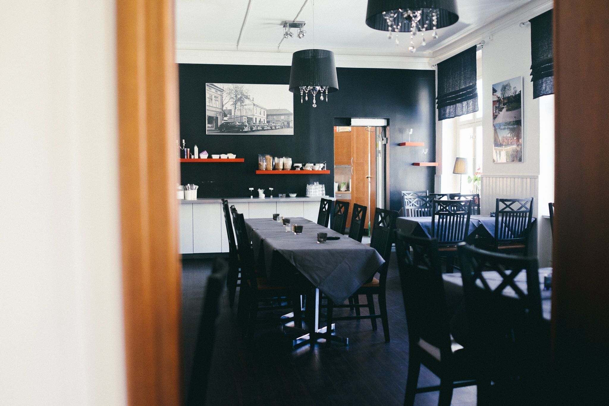 A dining area with a long table, chairs, and a chandelier. There are shelves with dishes and a window with curtains.