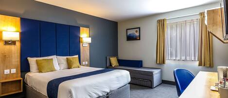 A hotel room with a large bed, a desk, and a chair. There is a window with curtains, a painting on the wall, and a lamp on the bedside table.