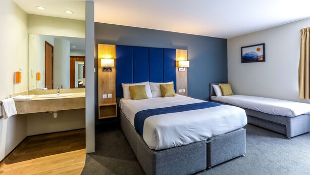 A hotel room with a large bed, a smaller bed, a bathroom with a sink and mirror, and a painting on the wall. - Days Inn by Wyndham Peterborough (Peterborough)
