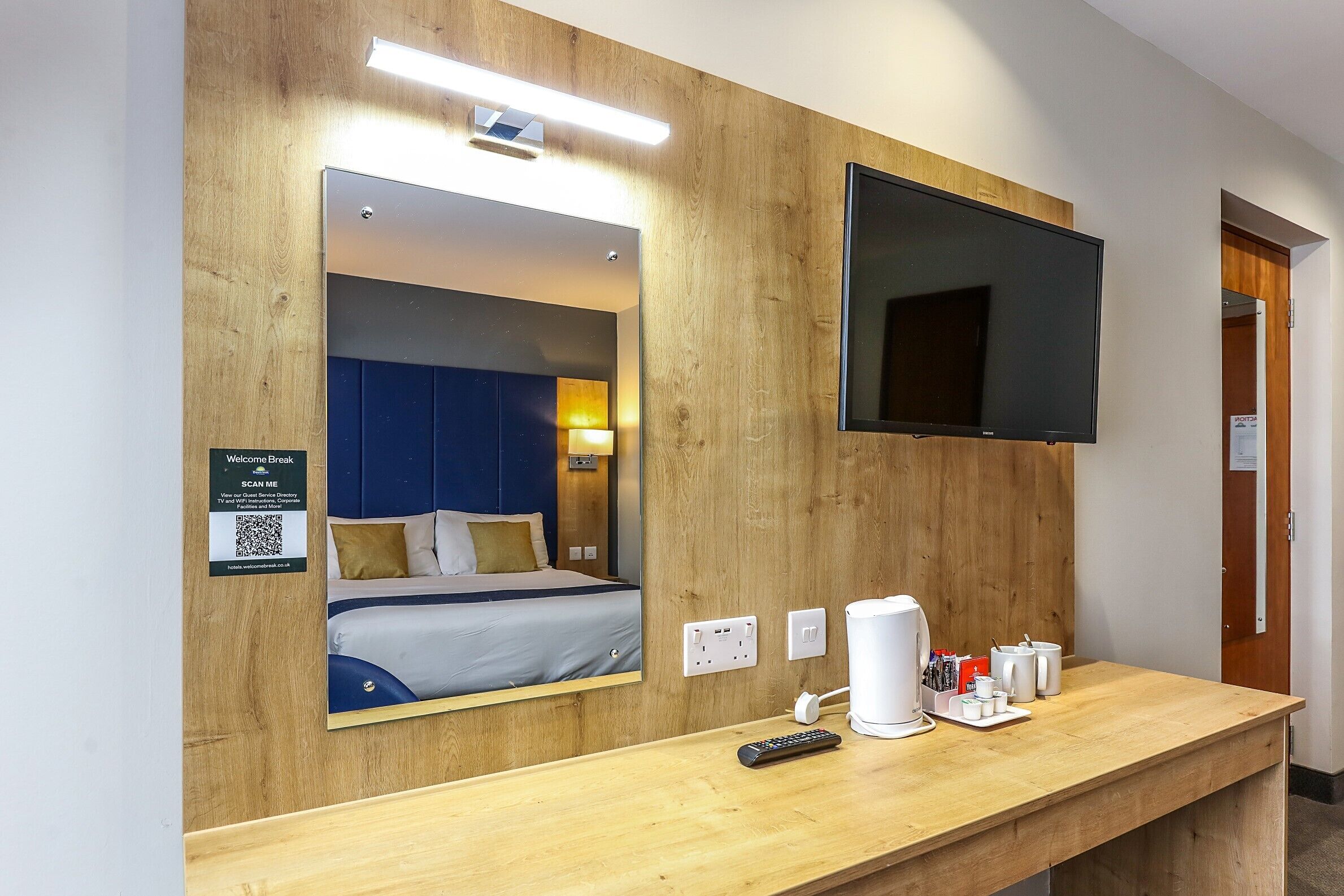 Habitación de hotel con cama, televisión, espejo, escritorio de madera con control remoto y estación de café.