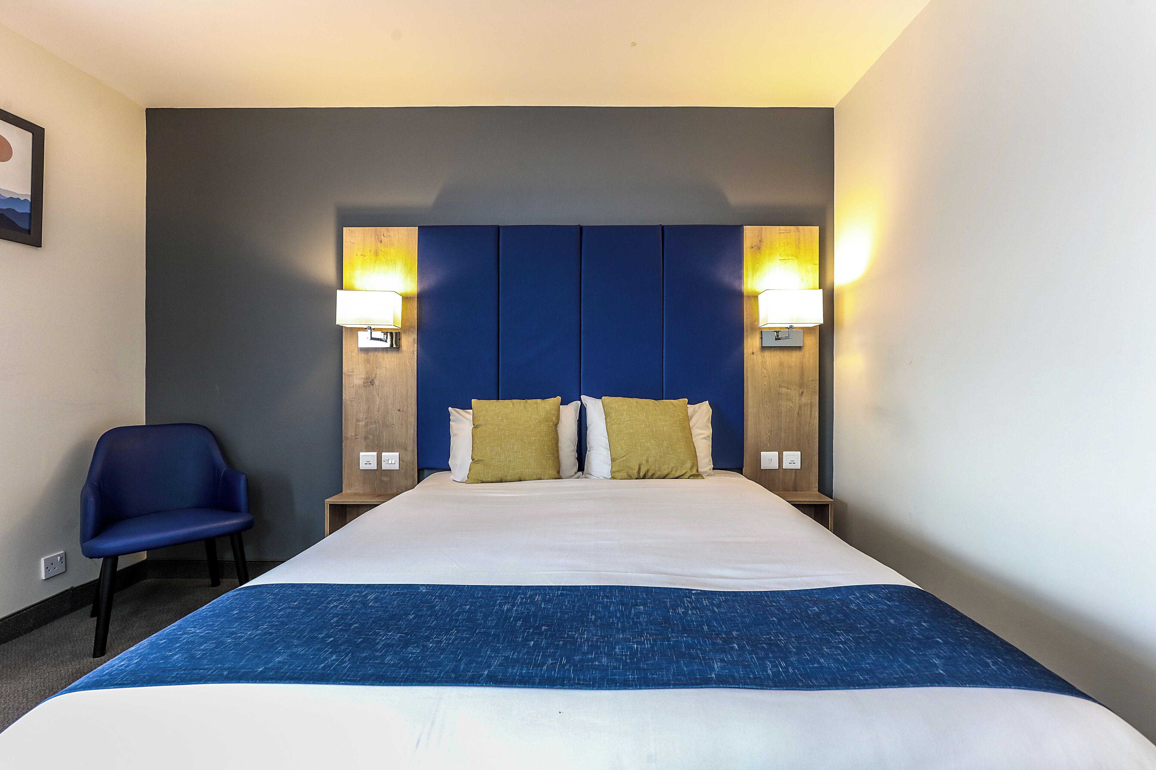 A hotel room with a large bed, two bedside lamps, a blue chair, and a framed picture on the wall.