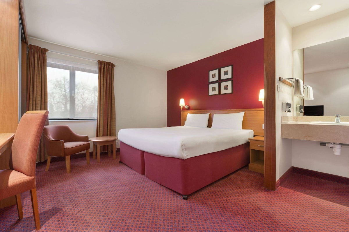 Habitación de hotel con cama, escritorio, silla, una mesita, espejo y ventana con cortinas.