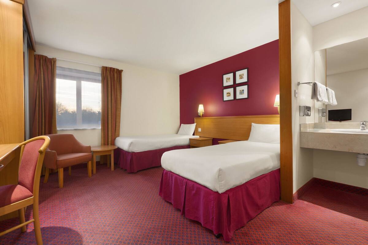 Habitación de hotel con dos camas, un escritorio, una silla, una mesita, un espejo y una ventana con cortinas.