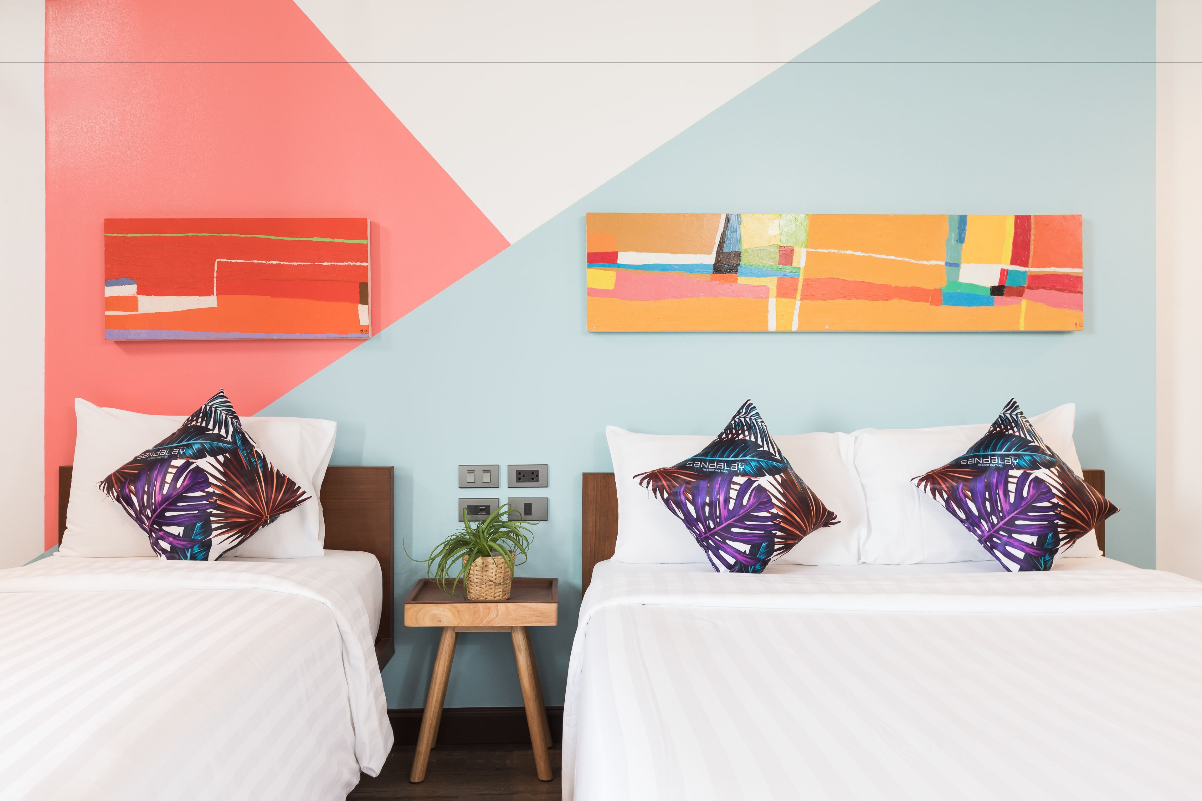 A hotel room with two beds, each with a white bedspread and a tropical leaf-patterned pillow. There is a small wooden nightstand with a plant and a wall-mounted light switch.