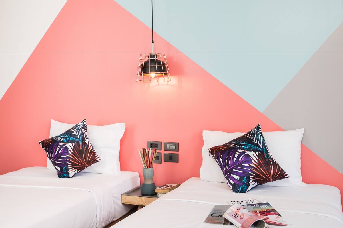 A bedroom with two beds, each with a white mattress and a colorful leaf-patterned pillow. A hanging light fixture is above the beds, and a magazine is on the floor.