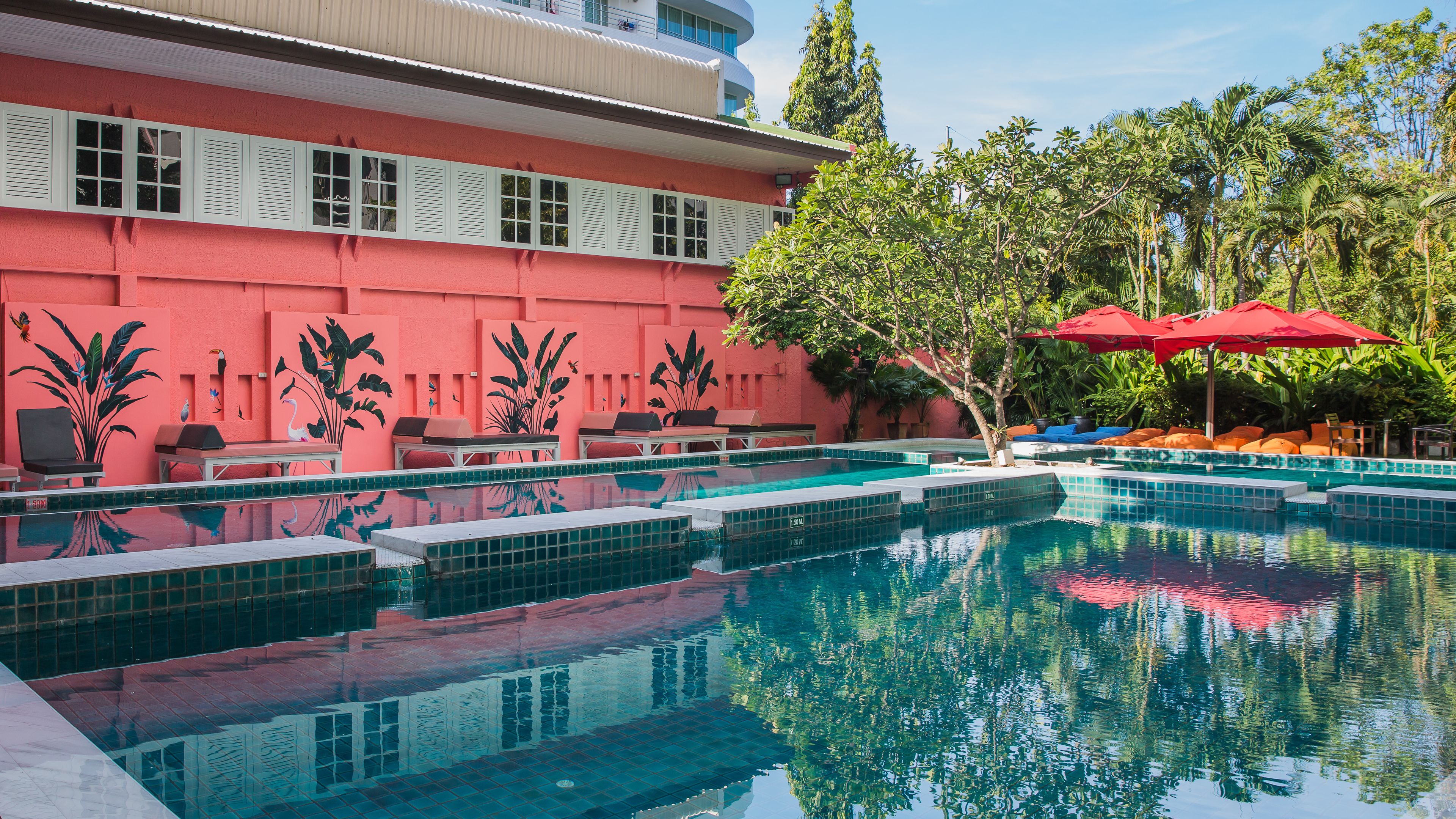 A swimming pool with lounge chairs and umbrellas, surrounded by a building with a red facade and tropical plants.
