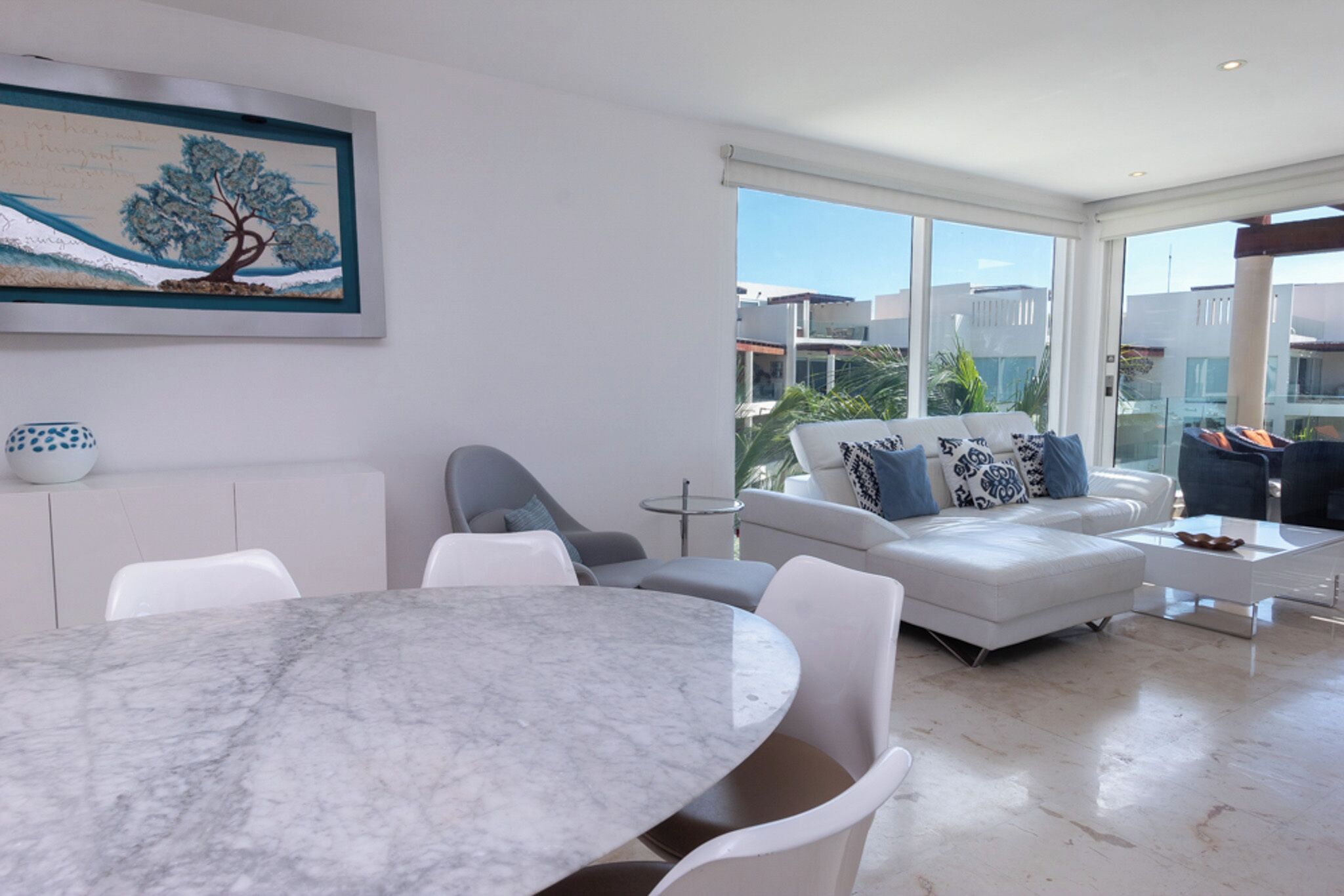 A modern living room with a marble table, white sofas, and a large window offering a view of buildings and greenery.