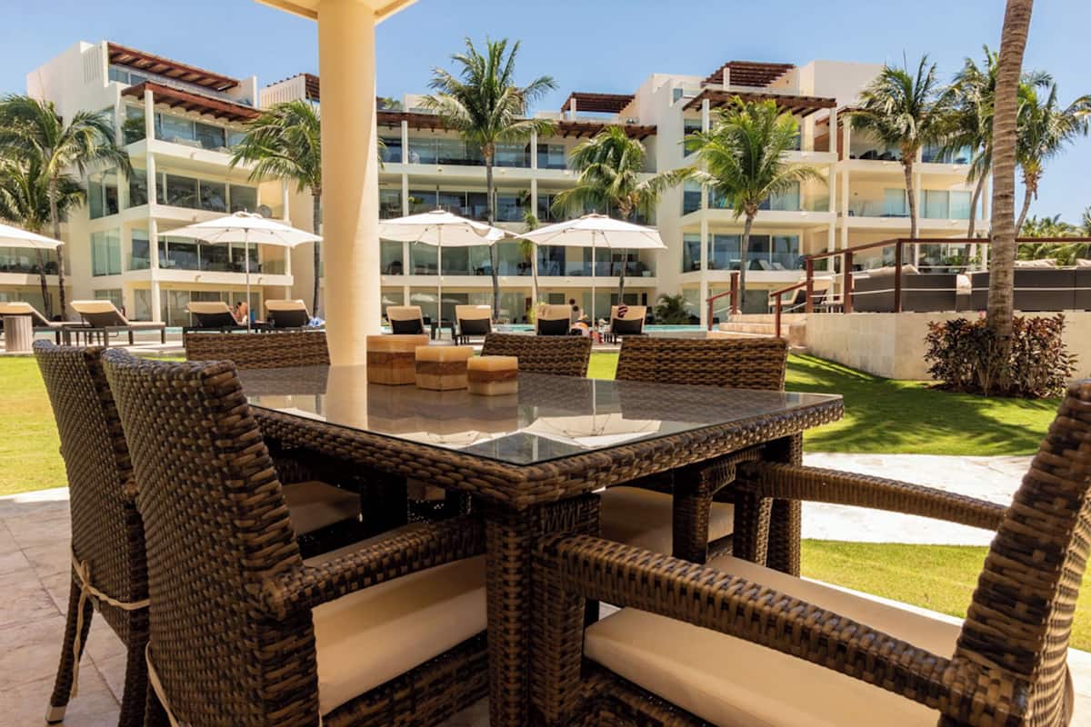 Un área de estar al aire libre con mobiliario de mimbre, una mesa con superficie de vidrio, sillones de descanso y palmeras, frente a un edificio con varios balcones.