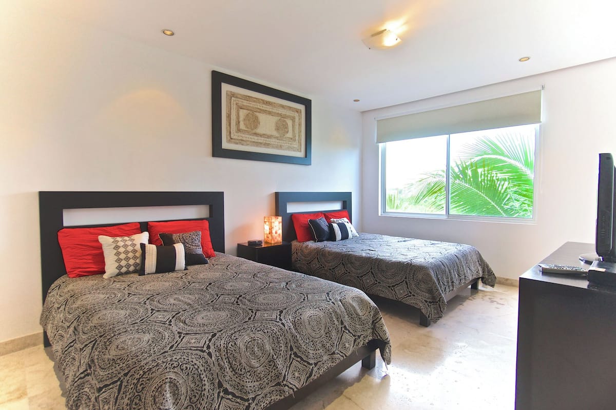 Habitación de hotel con dos camas, un ventanal con vista a palmeras, una obra de arte enmarcada en la pared y un escritorio con televisión.