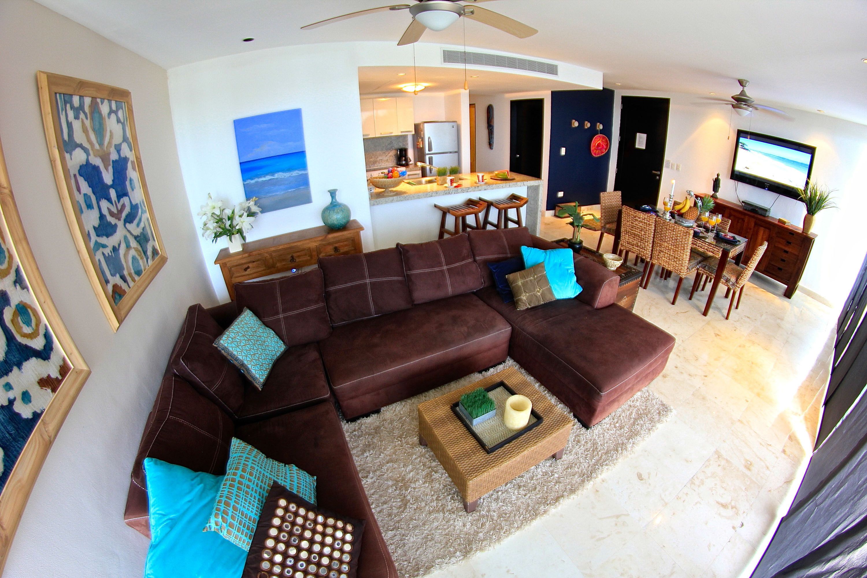 A modern living room with a sectional sofa, a coffee table, and a dining area with a TV and kitchen in the background.