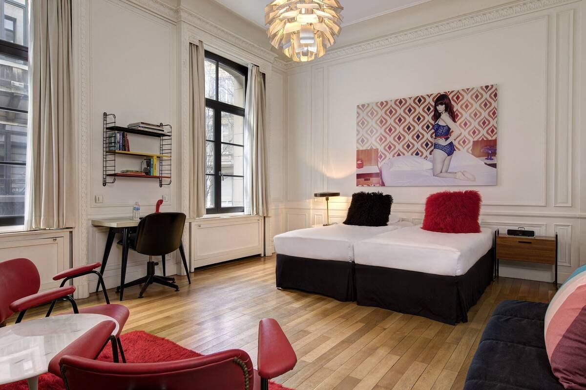 Ein modernes Hotelzimmer mit einem großen Bett, einem Schreibtisch und einem roten Sessel. An der Wand befinden sich ein Bücherregal und ein Gemälde.