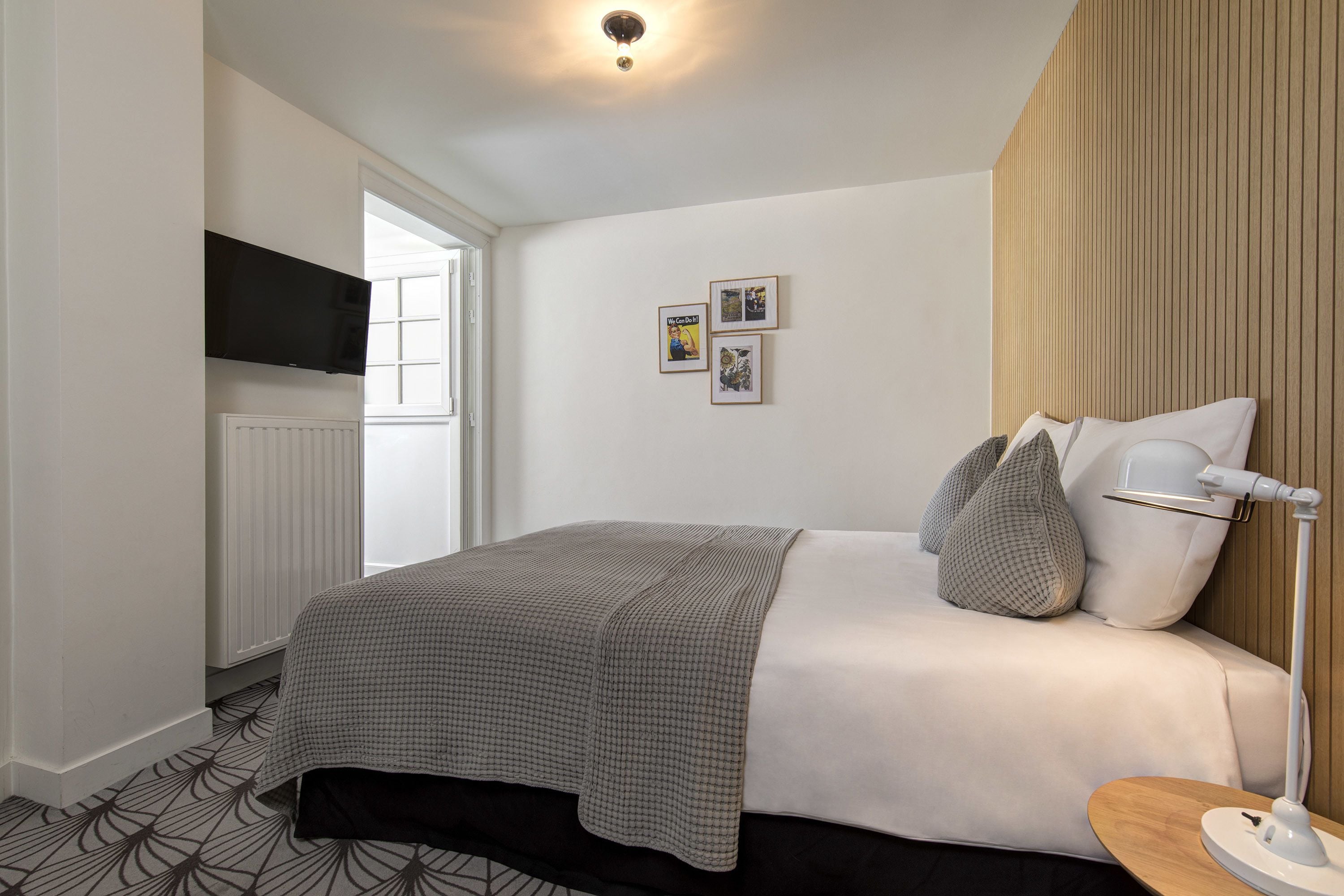 Un dormitorio moderno con cama, mesita de noche, lámpara, televisor montado en la pared y ventana.